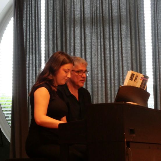 Eine Frau spielt Klavier, während ein älterer Mann zuschaut, mit einem Buch auf dem Klavier und grauen Vorhängen im Hintergrund.