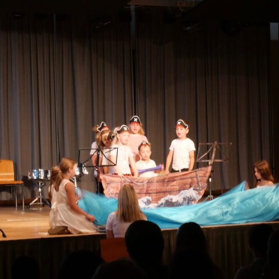 Eine Gruppe von Kindern, die als Piraten verkleidet sind, führt eine Aufführung auf der Bühne mit einem Boot als Requisite und Musikinstrumenten im Hintergrund durch.