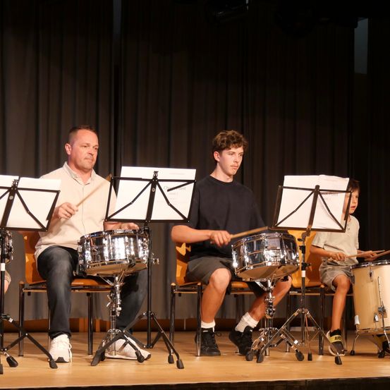 Drei Personen, darunter ein junger Junge, sitzen auf der Bühne mit Trommeln und Drumsticks, wahrscheinlich bei einer musikalischen Aufführung.