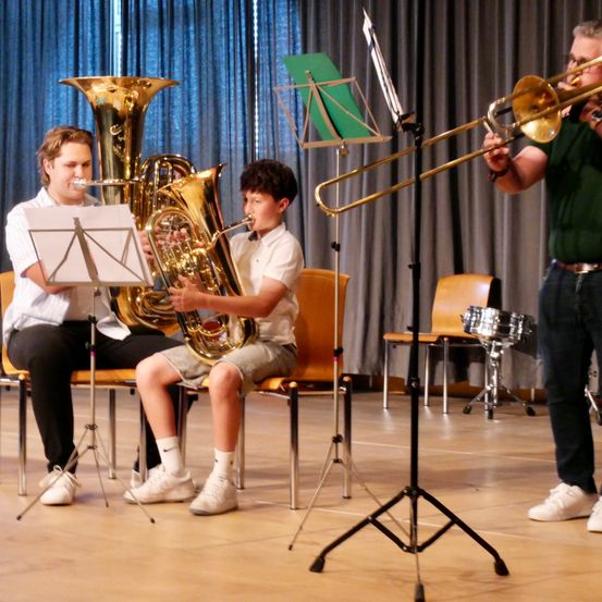 Drei Musiker spielen Instrumente in einem Raum. Einer spielt eine Posaune, ein anderer ein Tuba und der dritte spielt Schlagzeug. Sie sitzen auf Stühlen mit Notenständern vor ihnen.