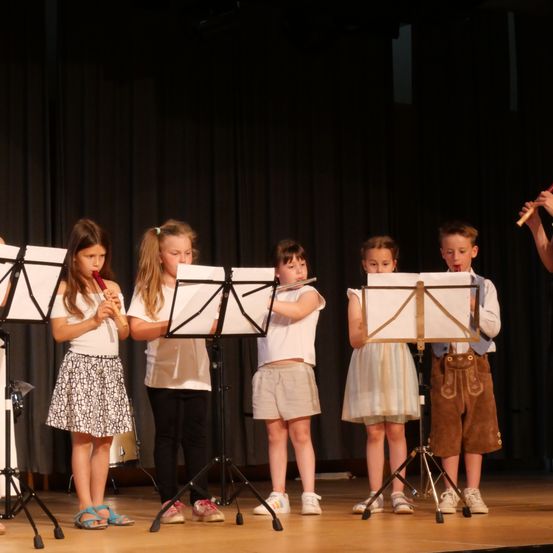 Eine Gruppe von Kindern in traditioneller Kleidung spielt Flöten auf einer Bühne mit Notenständern vor ihnen.