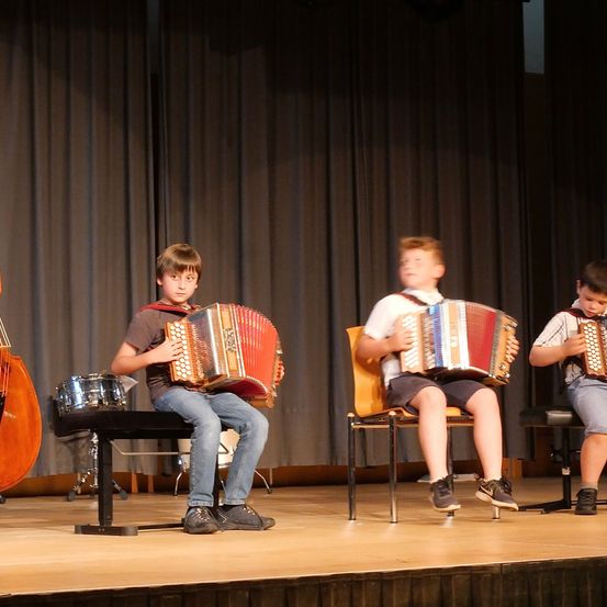 Drei junge Jungen spielen auf der Bühne, jeder ein Akkordeon. Der Junge auf der linken Seite sitzt auf einer Bank, während die anderen in Stühlen sitzen. Hinter ihnen sind ein Cello und ein Schlagzeug zu sehen.