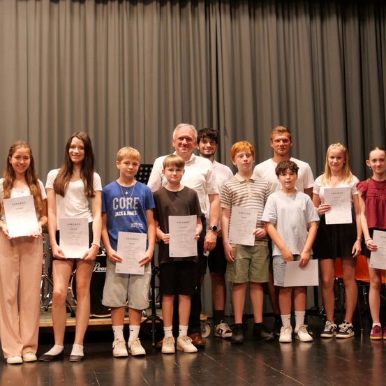 Eine Gruppe von Kindern und Erwachsenen steht auf einer Bühne und hält Urkunden in den Händen. Die Kinder tragen Freizeitkleidung und Turnschuhe. Die Erwachsenen tragen weiße Hemden und Turnschuhe. Ein grauer Vorhang ist im Hintergrund.