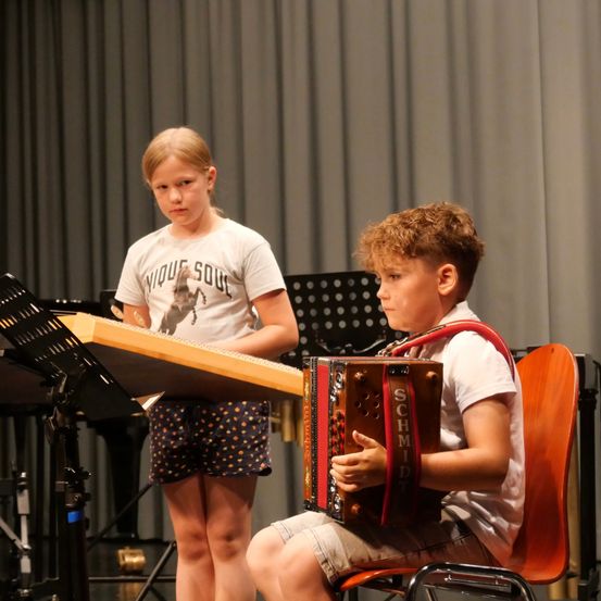 Zwei Kinder spielen Musikinstrumente in einem Studio. Ein Mädchen spielt ein hölzerne Xylophon, während ein Junge ein Akkordeon spielt.