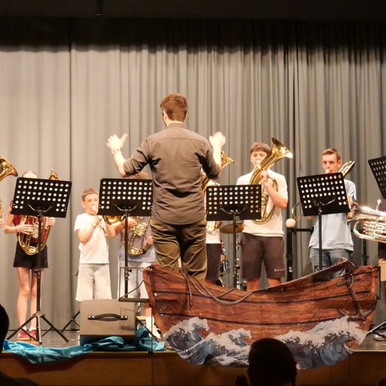 Ein Dirigent steht vor einer Gruppe junger Musiker, die auf der Bühne Blechblasinstrumente spielen, mit einem Bootsprop in der Vorderansicht.
