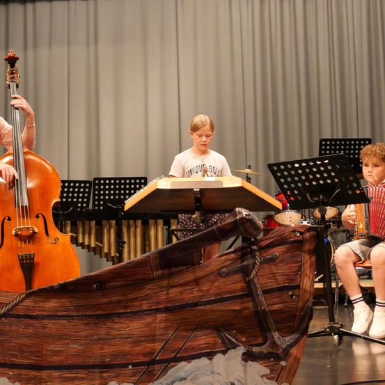 Ein junger Junge spielt Akkordeon, während ein anderer Cello und ein dritter Xylophon auf einer Bühne spielt, mit einem hölzernen Bootsprop in der Vorderansicht.