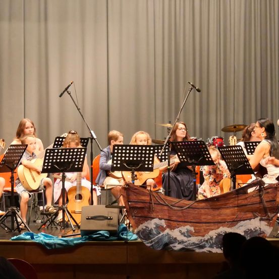 Eine Gruppe von Kindern und Erwachsenen spielt auf der Bühne mit Gitarren, Trommeln und einem großen Bootsprop.