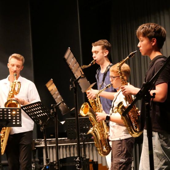 Vier junge Musiker, zwei Erwachsene und zwei Kinder, spielen Saxophon auf der Bühne mit Notenständern vor ihnen.