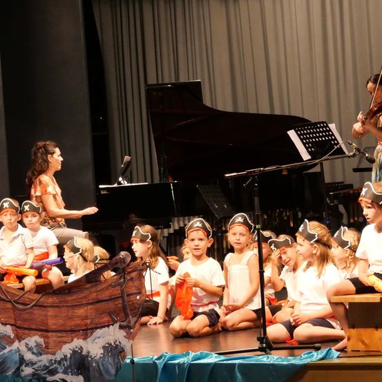 Eine Gruppe von Kindern, die als Piraten verkleidet sind, tritt mit einer Frau und einer Violinistin auf der Bühne auf. Ein Klavier befindet sich hinter ihnen.