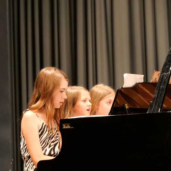 Ein junges Mädchen spielt Klavier, während zwei andere Mädchen aufmerksam zuschauen, in einem schwach beleuchteten Raum.