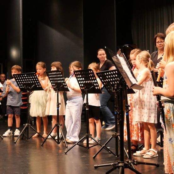 Eine Gruppe von Kindern in einem Schulstück, die Flöten halten und vor Musikständen auf der Bühne stehen.