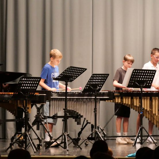 Drei junge Jungen spielen Xylophon auf der Bühne vor einem Publikum. Jeder Junge konzentriert sich auf sein Instrument, mit Notenständern und Noten vor ihnen.