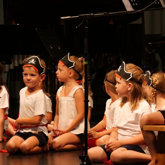 Eine Gruppe von Kindern in Piratenhüten sitzt auf dem Boden, wahrscheinlich bei einer Aufführung oder einem Spiel. Sie tragen weiße Hemden und Shorts.