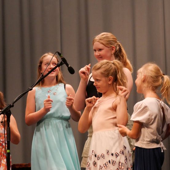 Vier junge Mädchen stehen auf einer Bühne, zwei von ihnen halten Mikrofone und singen. Das Mädchen in der Mitte gestikuliert mit ihren Händen. Hinter ihnen ist ein grauer Vorhang.