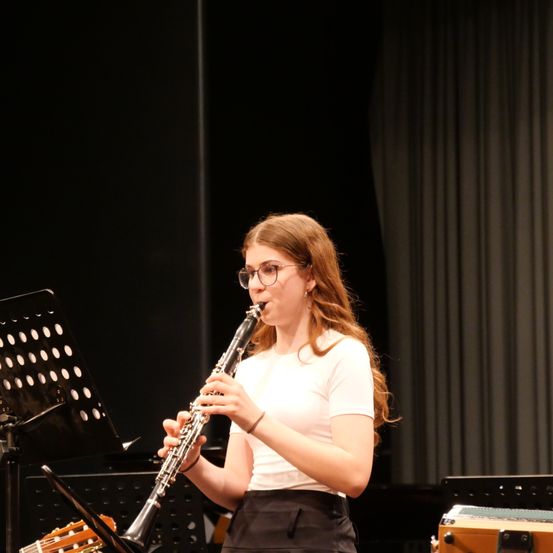 Eine junge Frau mit Brille spielt Klarinette auf der Bühne, mit einem Notenständer und einer Gitarre im Hintergrund.