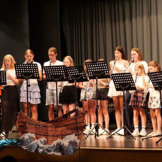Eine Gruppe junger Musiker, in lässiger Kleidung gekleidet, spielt auf der Bühne mit ihren Klarinetten. Die Bühne ist mit einem Hintergrund aus schwarzen Vorhängen und einem großen Bootsprop in der Vorderansicht ausgestattet.