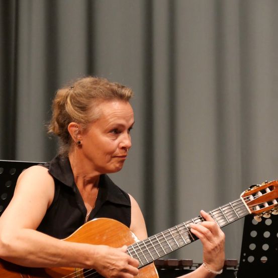 Eine Frau mit kurzen Haaren spielt Gitarre und schaut zur Seite. Sie trägt ein schwarzes ärmelloses Oberteil und hat einen ernsten Gesichtsausdruck.