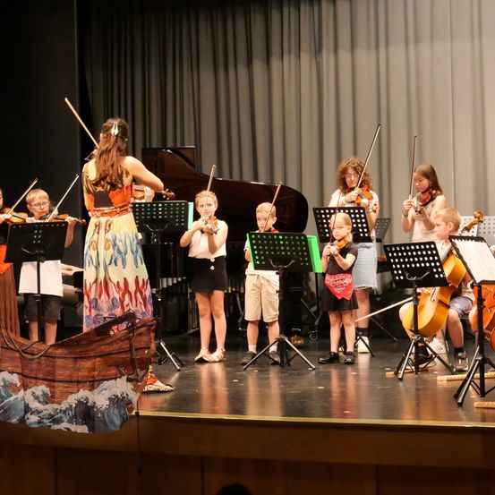 Eine Gruppe von Kindern spielt Violinen auf der Bühne mit einem Schiffshintergrund, begleitet von einem Pianisten.