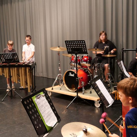 Eine Gruppe von Kindern und einer erwachsenen Frau spielt verschiedene Musikinstrumente, darunter Trommeln, Xylophon und Becken, in einem Klassenzimmer.