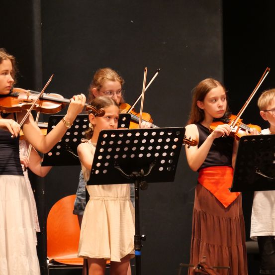 Eine Gruppe junger Mädchen spielt Violinen auf der Bühne, mit Notenständern vor ihnen. Sie sind in verschiedenfarbigen Kleidern gekleidet.
