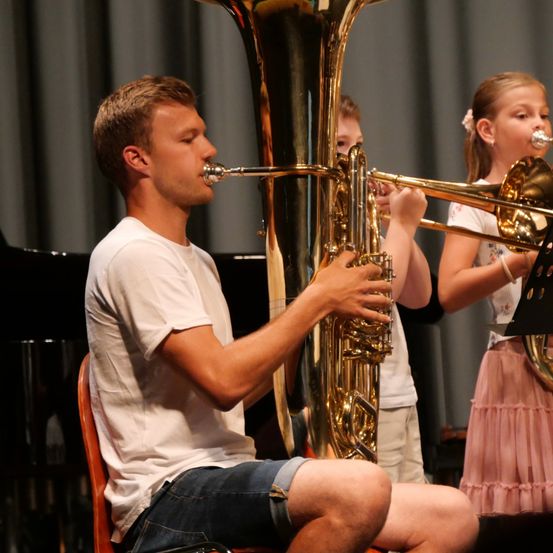 Ein junger Mann spielt eine Tuba im Sitzen, begleitet von zwei Kindern, die Trompeten spielen, in einer Aufführungssituation.