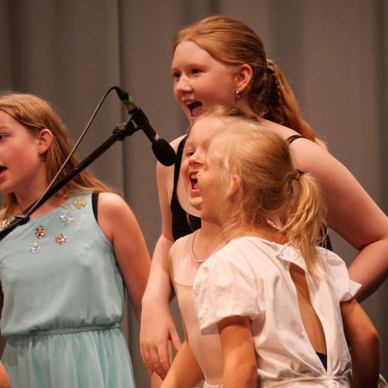 Drei junge Mädchen singen in ein Mikrofon, eine in einem blauen Kleid und zwei weitere in weißen Kleidern.