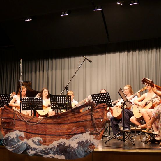 Eine Gruppe von Musikern tritt auf der Bühne auf, einige sitzen auf Stühlen und andere auf einem bootförmigen Requisit. Sie spielen Gitarre und singen.