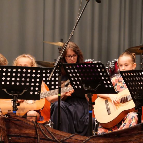 Drei junge Mädchen spielen Gitarren auf der Bühne mit einer Frau im Hintergrund, begleitet von Trommeln und Becken.