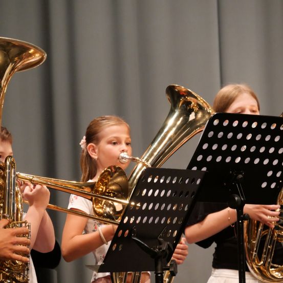 Drei Kinder spielen Blechblasinstrumente, darunter Posaunen und Tuben, in einem Schulmusikraum. Sie konzentrieren sich auf ihre Notenständer vor ihnen.