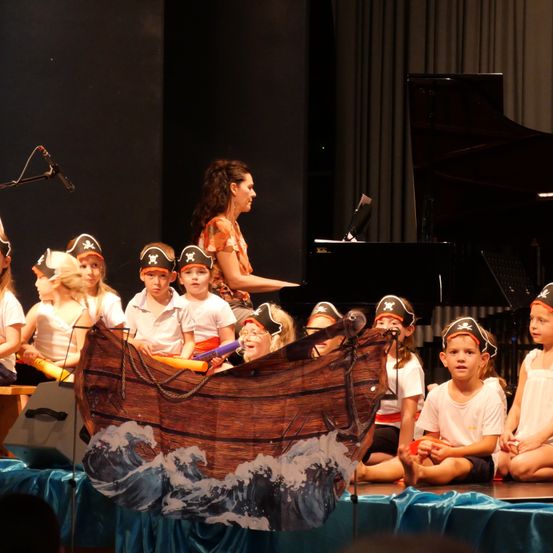 Eine Gruppe von Kindern in Piratenhüten führt mit einer Frau, die Klavier spielt, auf der Bühne auf.
