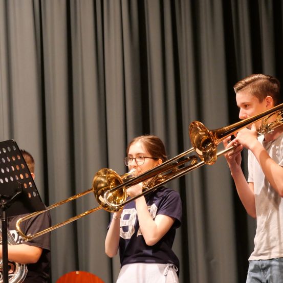 Zwei Schüler spielen Posaunen auf der Bühne mit einem Vorhang im Hintergrund. Das Mädchen trägt eine Brille und ein blaues Shirt, während der Junge ein weißes Shirt trägt.