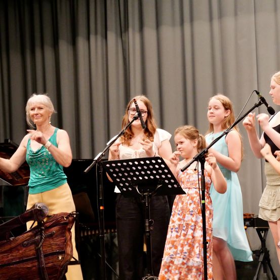 Eine Gruppe von Frauen und Mädchen tritt mit Musikinstrumenten auf der Bühne auf. Die Frau im grünen Oberteil singt, während die anderen Instrumente spielen.