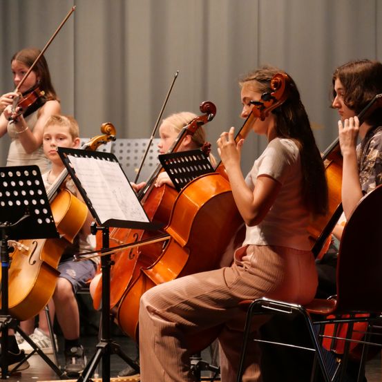 Eine Gruppe junger Musiker, darunter Mädchen und Jungen, spielen Cellos und Violinen auf der Bühne während einer Aufführung.