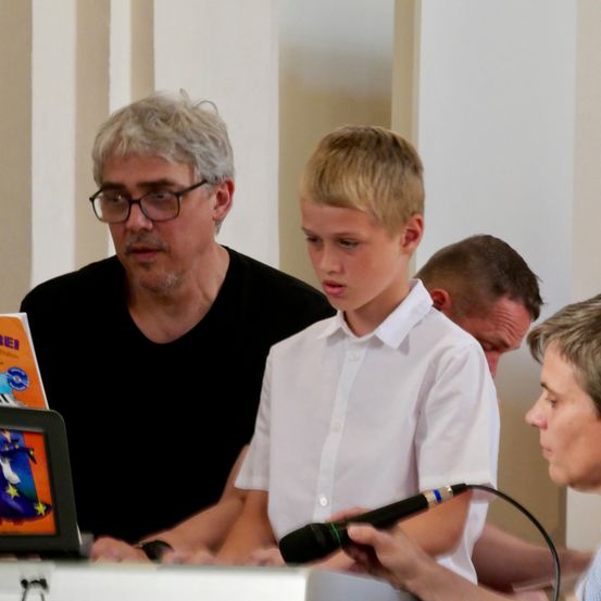 Ein Junge mit einem Mikrofon steht vor einem Schreibtisch, hinter ihm steht ein älterer Mann in einem schwarzen T-Shirt. Ein weiterer Mann sitzt hinter ihnen, und ein Buch liegt auf dem Schreibtisch.