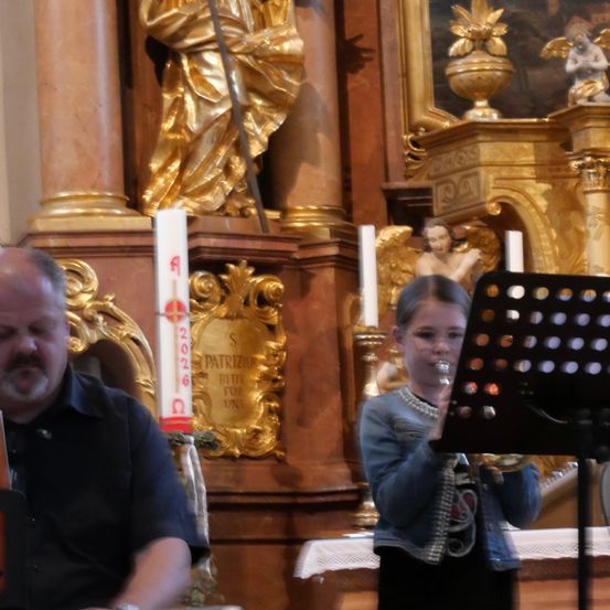 Ein junges Mädchen spielt Trompete in einer Kirche, neben ihr sitzt ein Mann. Dahinter steht eine goldene Statue und eine Kerze.