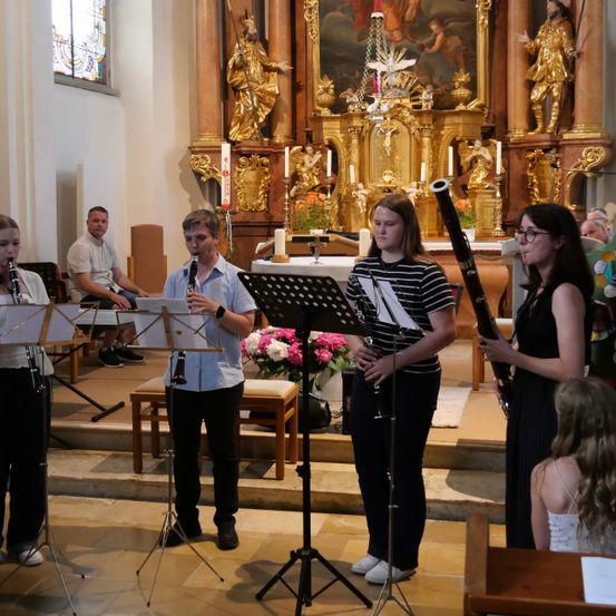 Eine Gruppe junger Musiker tritt auf der Bühne in einer Kirche auf, wobei ein Mann Klarinette und drei Frauen Fagott spielen. Hinter ihnen steht ein geschmückter Altar mit Kerzen und einem Gemälde.