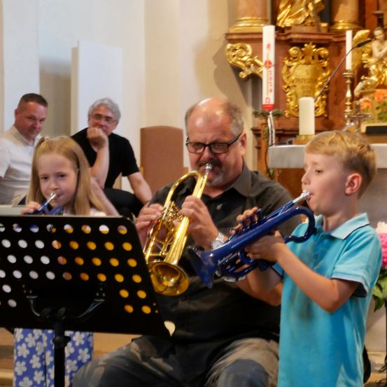 Ein Mann und zwei Kinder spielen Trompeten in einer Kirche. Der Mann sitzt, während die Kinder stehen. Hinter ihnen sitzen einige Leute auf Stühlen.