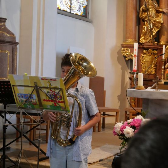 Ein junger Junge spielt eine Blechposaune in einer Kirche, mit einem Notenständer vor ihm und einer goldenen Statue dahinter.