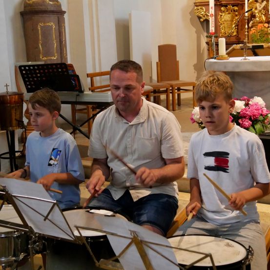 Ein Mann und zwei Jungen spielen in einer Kirche Trommeln. Der Mann sitzt und die Jungen stehen. Musikinstrumente sind um sie herum aufgestellt.