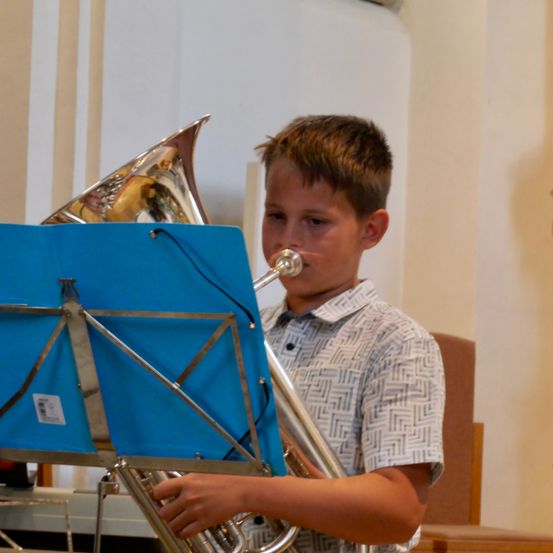 Ein junger Junge spielt eine silberne Tuba mit einem blauen Notenständer in einem Raum.