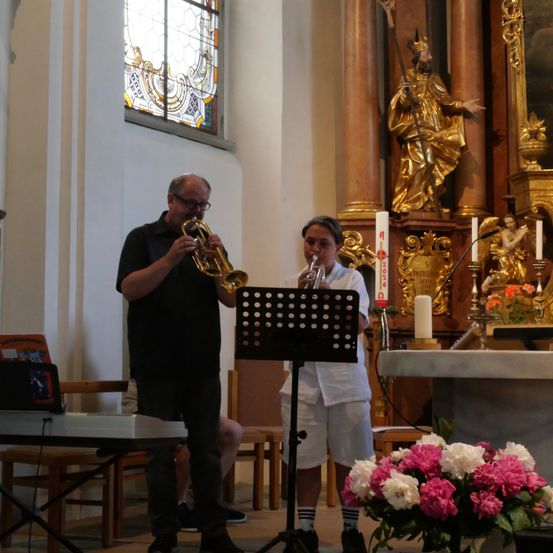 Zwei Personen spielen Trompeten in einer Kirche, mit einer goldenen Statue im Hintergrund. Ein Blumenstrauß steht auf dem Altar.