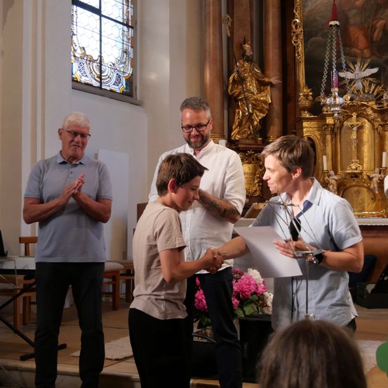 Ein junger Junge gibt einer Frau in einer Kirche die Hand, während ein Mann hinter ihnen klatscht. Eine goldene Statue und ein Gemälde sind im Hintergrund.