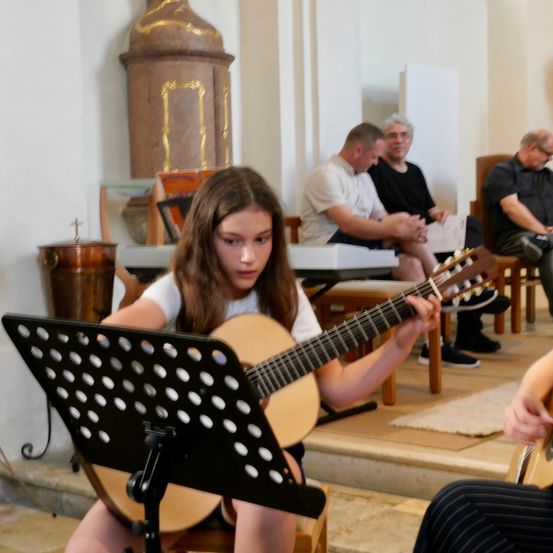 Ein junges Mädchen spielt Gitarre in einer Kirche, während drei Männer im Hintergrund zusehen.