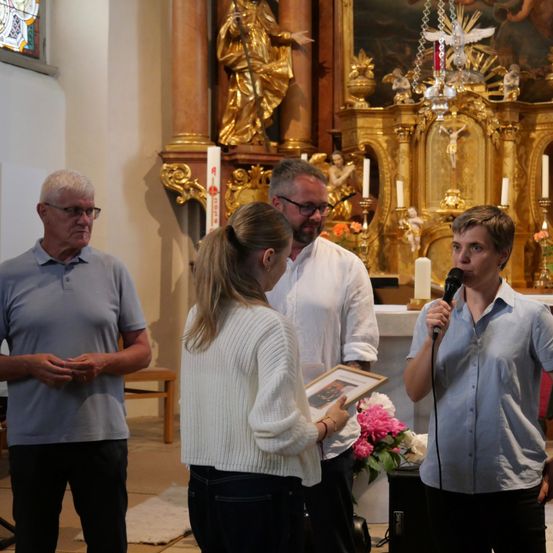 Vier Menschen stehen in einer Kirche, zwei Männer und zwei Frauen, einer hält ein gerahmtes Bild, der andere ein Mikrofon.