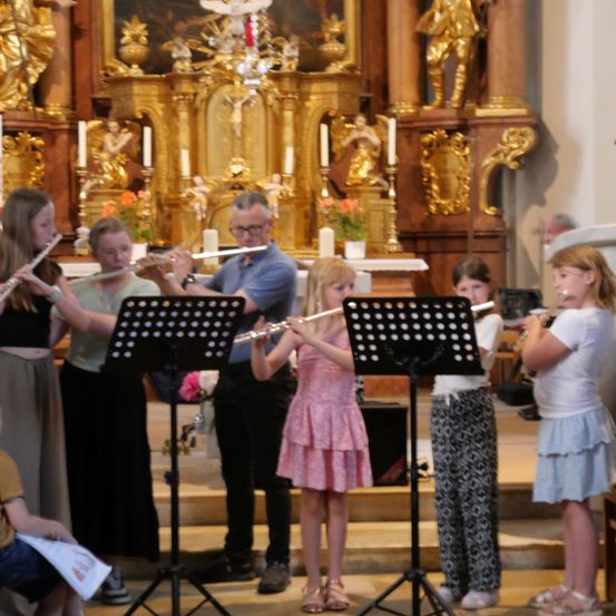 Eine Gruppe von Kindern und einem Erwachsenen spielen Flöten vor einem Kirchenaltar. Die Kinder sind in Freizeitkleidung gekleidet, und der Erwachsene trägt eine Brille.
