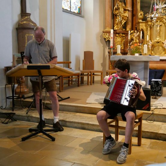 Ein älterer Mann und ein junger Junge spielen Musikinstrumente in einer Kirche. Der Mann spielt ein Akkordeon, während der Junge ein kleines Akkordeon spielt.