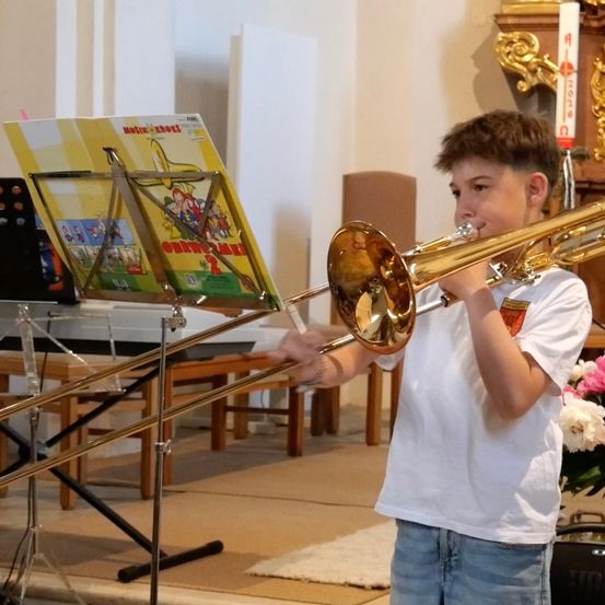 Ein junger Junge spielt eine Posaune in einer Kirche, mit einem Notenbuch vor ihm.