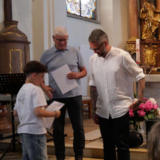 Drei Männer und ein Junge stehen in einer Kirche und halten Papiere in der Hand. Ein Mann trägt eine Brille, ein anderer hat Tattoos. Im Hintergrund steht eine Statue und Blumen.