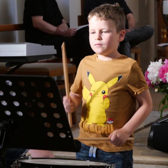 Ein junger Junge mit einem Pikachu-T-Shirt spielt mit einem Drumstick in der Hand die Trommel. Dahinter sitzt eine Person auf einem Stuhl.