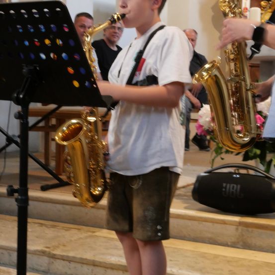 Ein junger Junge spielt in einer Kirche ein Saxophon, begleitet von einem anderen Musiker. Ein Notenständer und ein JBL-Lautsprecher sind in der Nähe.